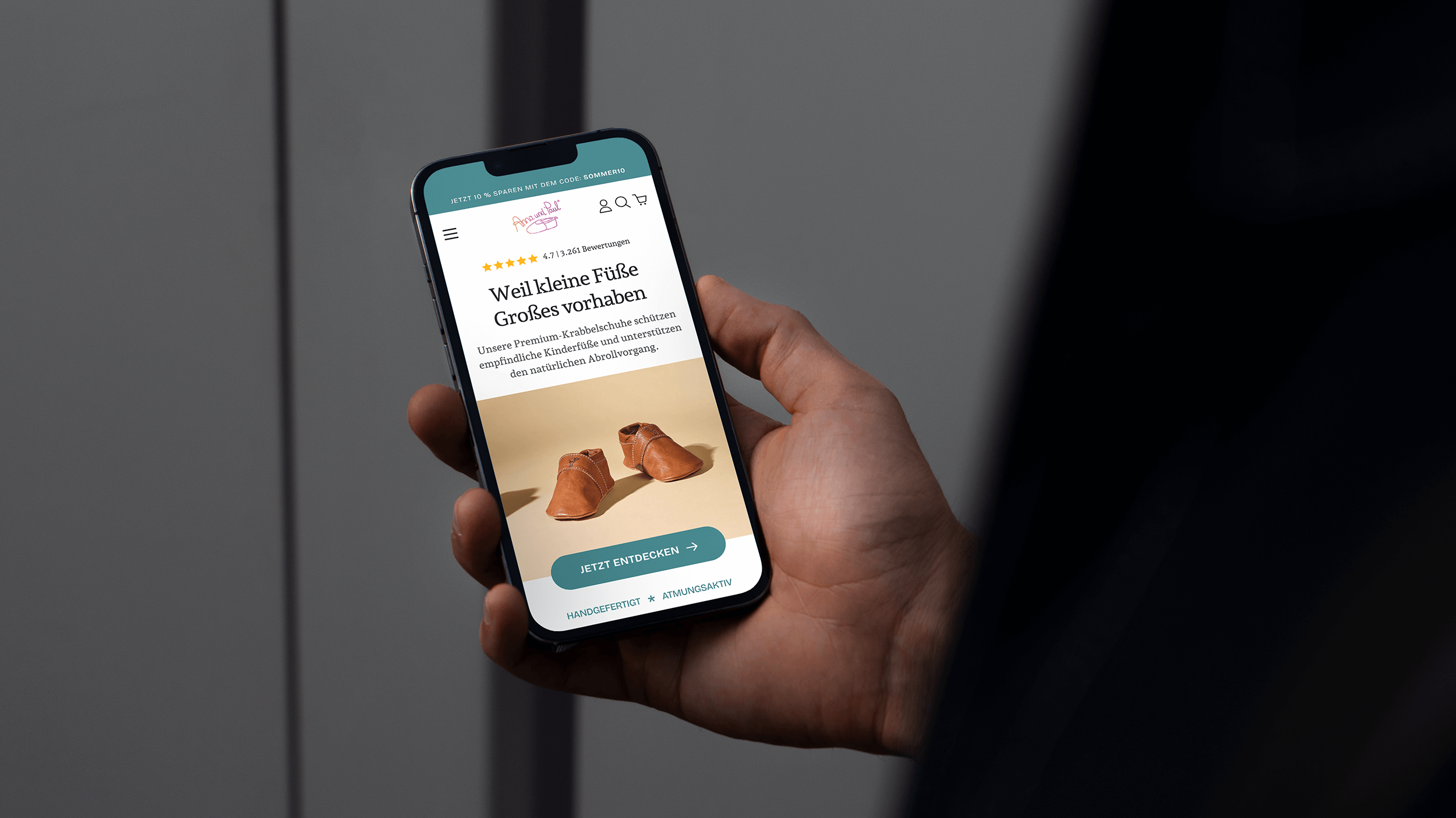 Unser Inhaber Tim steht vor einer Holzkommode, auf der ein runder Spiegel steht. Er blickt in die Kamera und hat ein zugeklapptes MacBook unter dem Arm.